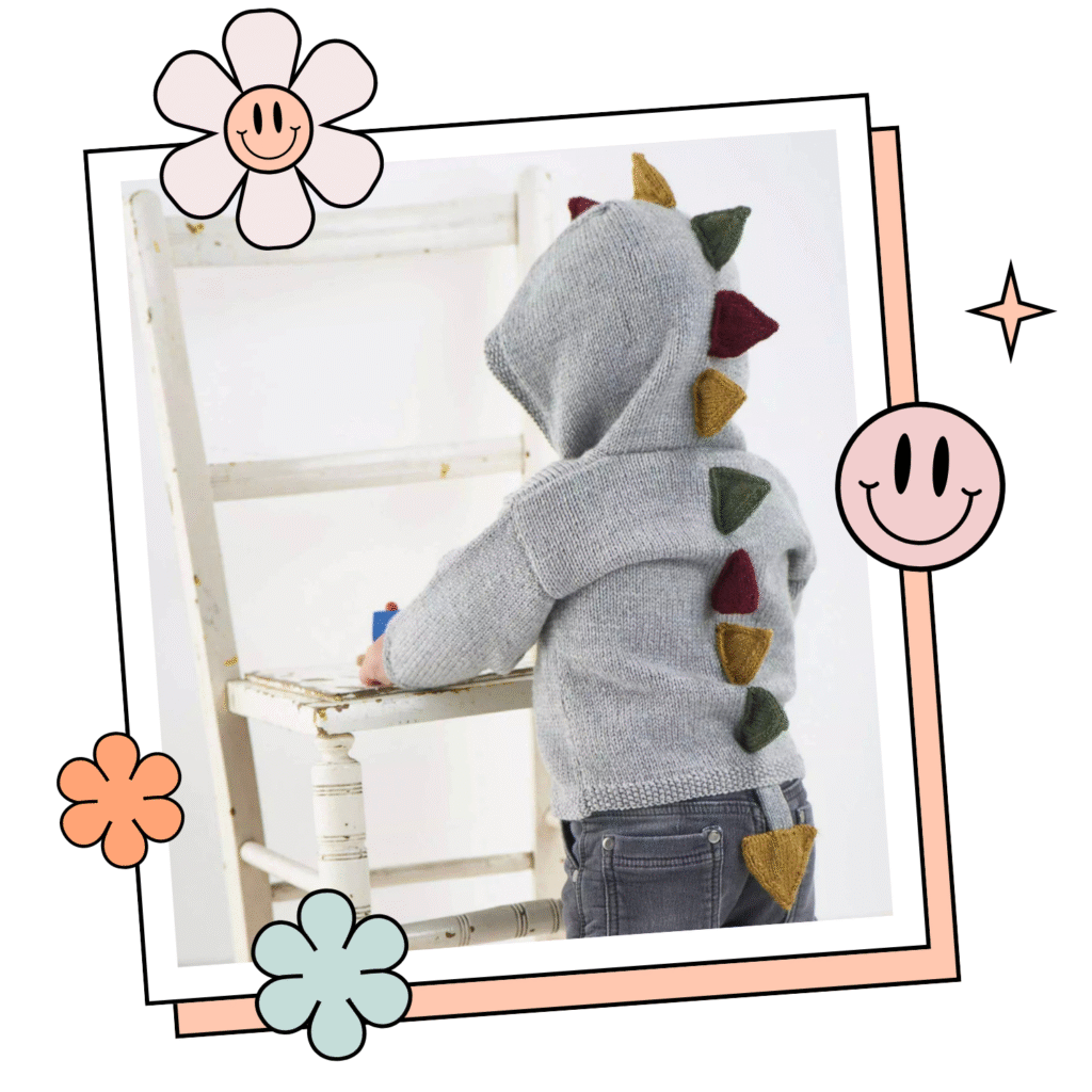 toddler wearing jane burns dinosaur explorer hooded cardigan knitting pattern. It is a light grey hooded cardigan with knitted spikes down the back of the hood and body. there is a tail at the centre back bottom of the cardigan.