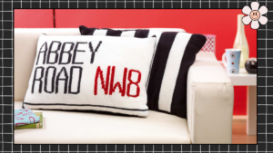 Two knitted cushion covers on a white sofa — Abbey Road NW8 street sign cushion in black, white and red, alongside a black and white striped zebra crossing cushion, knitting pattern by Jane Burns